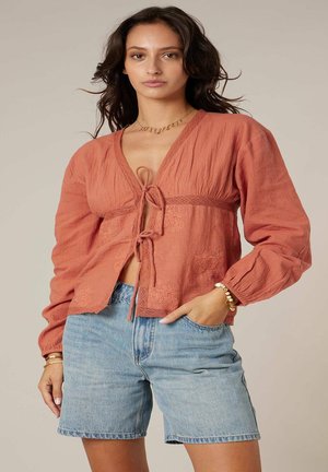 Femme portant un chemisier à manches longues couleur rouille avec des liens à l'avant et un short en denim bleu clair, debout avec une main dans la poche.