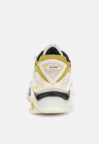Sneaker chunky con una parte superiore bianca, accenti neri e gialli, texture in rete e suede, con un logo triangolare sul tallone.