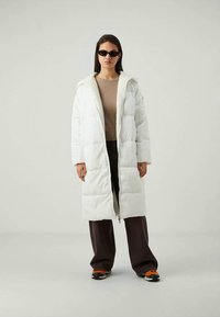 Kvindelig i en lang hvid pufferjakke over en beige sweater, mørke vide bukser, orange sneakers og sorte solbriller, stående mod en ensfarvet baggrund.