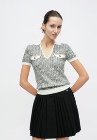 Haut à manches courtes texturé gris et blanc avec un col en V, comportant deux poches poitrine blanches avec des accents dorés ; associé à une jupe plissée noire.