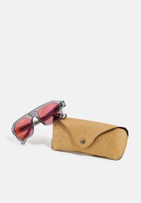 Occhiali da sole grigi trasparenti con lenti rosa accanto a una custodia in suede beige con chiusura a scatto e logo impresso.