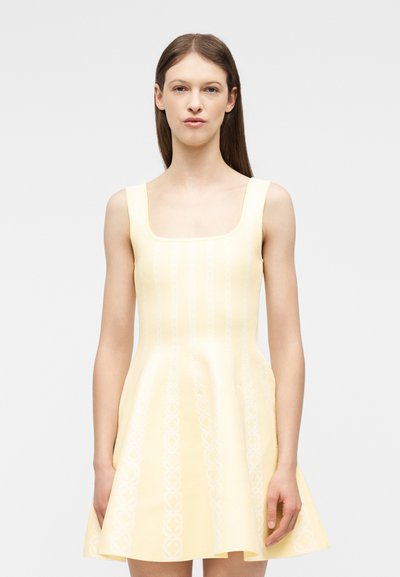 Γυναίκα με μακριά καστανά μαλλιά, φορώντας ένα αμάνικο ανοιχτό κίτρινο φόρεμα με τετράγωνη λαιμόκοψη και φαρδιά φούστα, στέκεται μπροστά σε λευκό φόντο.