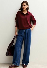 Polo shirt bordeaux a maniche lunghe con colletto, abbinato a jeans in denim blu a vita alta e gamba larga. Sneakers marroni e una borsa scura completano il look.
