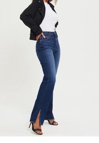 Jeans azul oscuro de cintura alta con aberturas laterales en el dobladillo, combinados con una blusa blanca de canalé y una chaqueta negra corta.