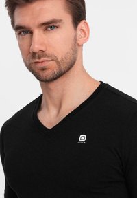 Černé, přiléhavé triko s výstřihem do V vyrobené z hladkého materiálu, které na levé straně hrudi má malé bílé logo. Design je jednoduchý a elegantní.