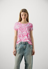 T-shirt rosa a maniche corte con un intricato motivo floreale bianco. Il materiale è morbido; scollatura rotonda. Indossato con jeans azzurri chiari, confortevoli e larghi.