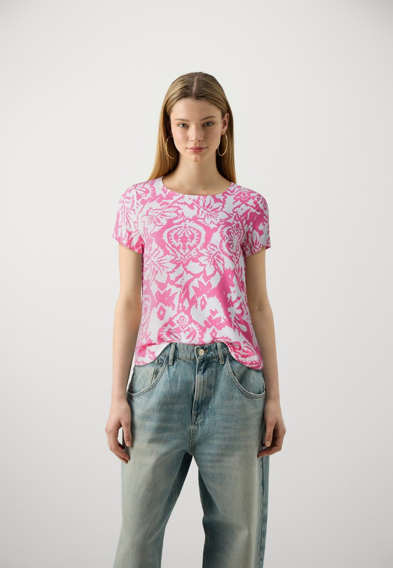 T-shirt rosa a maniche corte con un intricato motivo floreale bianco. Il materiale è morbido; scollatura rotonda. Indossato con jeans azzurri chiari, confortevoli e larghi.