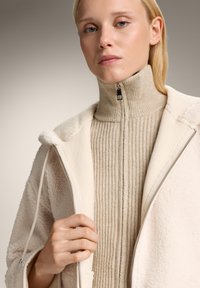 Giacca di shearling color crema parzialmente zipata su un dolcevita ribbed beige, con colletto alto e un sottile dettaglio con zip.