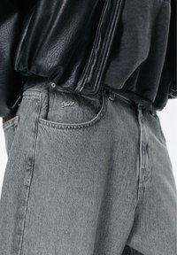 Graue Jeans aus Denim mit lockerem Schnitt, versehen mit einem Logo auf der vorderen Tasche, kombiniert mit einer schwarzen Lederjacke mit Reißverschluss.