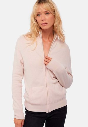 Femme blonde portant un sweat à capuche rose clair avec des poches à l'avant, à moitié zippé, et un pantalon noir sur fond uni.