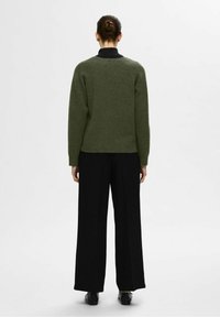 Maglione verde in lana con finitura testurizzata, vestibilità ampia e colletto alto, abbinato a pantaloni neri a gamba larga e scarpe nere.