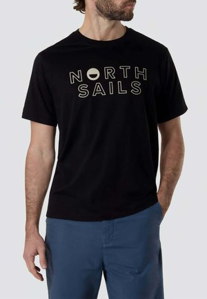 Uomo che indossa una maglietta nera a maniche corte con la scritta "NORTH SAILS" e pantaloni blu, mani rilassate, guarda di lato.