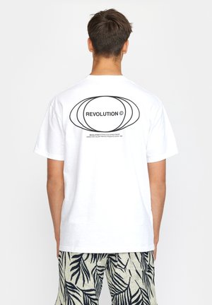 Uomo che indossa una maglietta bianca con motivo ovale sovrapposto e la scritta "REVOLUTION ©" sulla schiena, abbinata a pantaloncini neri e bianchi con fantasia a foglie.