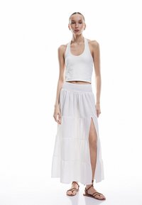 Top corto blanco sin mangas con un cuello alto, combinado con una falda larga blanca de volantes que presenta una abertura lateral, tela texturizada y cintura elástica.