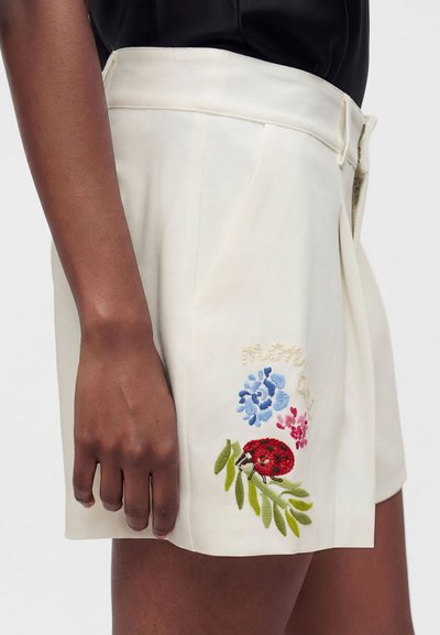 Personne portant un short crème avec une broderie florale colorée et une coccinelle rouge près de l'ourlet, assorti d'un haut noir.