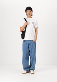 Bílý tričko s grafickým potiskem, volné modré džínové kalhoty a bílé tenisky s červenými akcenty. Černá crossbody taška. Jednoduchý, ležérní styl.