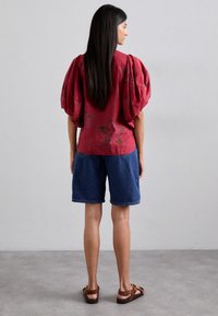 Punainen kukkakuosisella puserolla, jossa on puffihihat, yhdistettynä sinisiin farkkushortseihin. Malli seisoo ruskeissa sandaaleissa, nähtynä takaa.