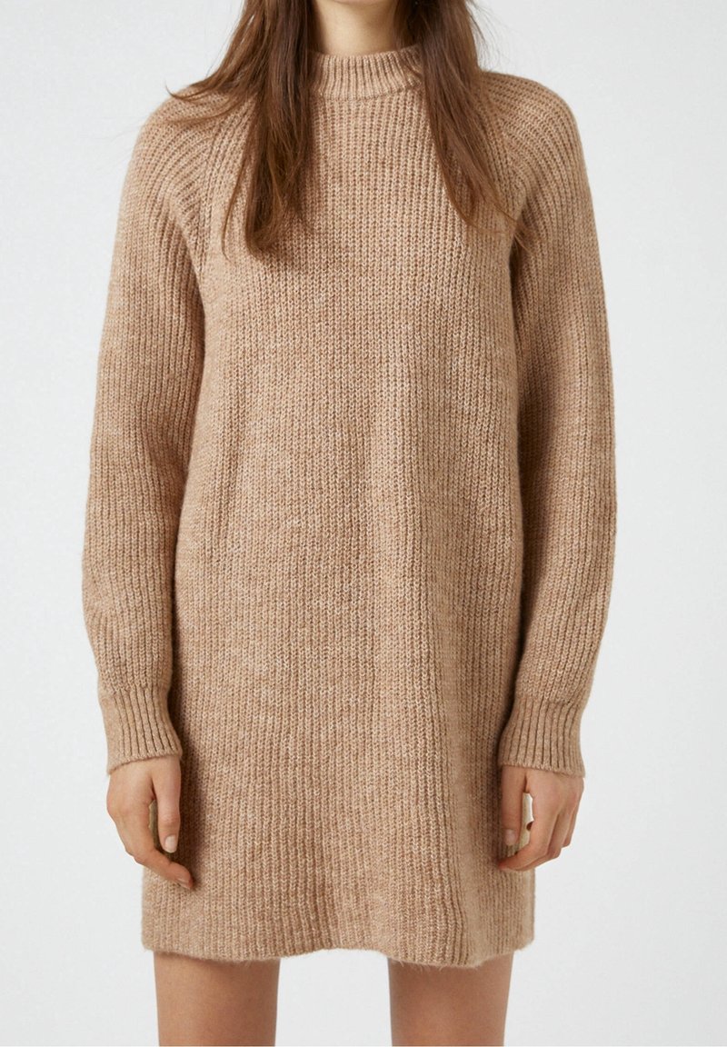 Femme portant une robe-pull en maille côtelée beige à manches longues avec un col montant, debout devant un fond uni.