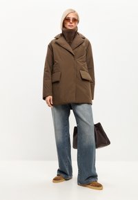 Abrigo acolchado marrón con cuello de chal y dos bolsillos frontales. Combinado con jeans de pierna ancha azules y calzado marrón, sosteniendo un bolso oscuro.