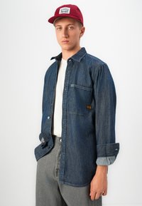 Young man wearing dark denim shirt over white tee, gray pants, and red cap, standing against a plain white background.