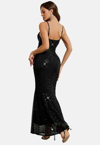 Frau in einem enganliegenden schwarzen Paillettenabendkleid mit Spaghettiträgern, barfuß stehend und über die Schulter blickend, vor einem schlichten Hintergrund.