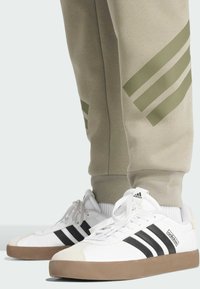 Witte sportschoenen met zwarte strepen en een beige rubberen zool, gecombineerd met lichtgrijze joggingbroeken met groene diagonale strepen.