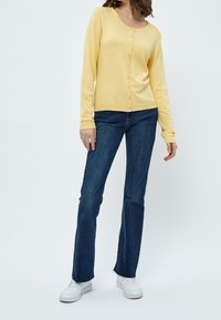 Gul långärmad cardigan med knäppning, ribbade ärmslut och nederkant, tillsammans med mörkblå utsvängda jeans och vita sneakers.