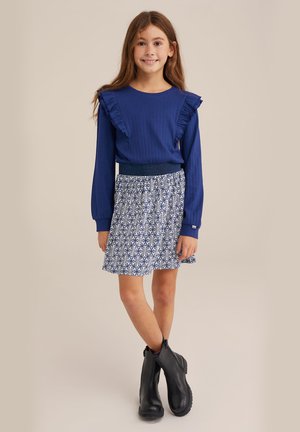 Blaue, gerippte Langarmbluse mit Rüschendetails, kombiniert mit einem blütenmuster-geprägten weißen und blauen Rock und schwarzen Knöchelstiefeln.