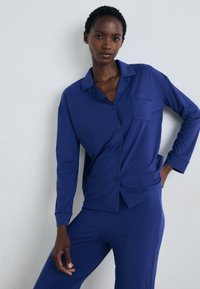 Mėlynas ilgomis rankovėmis pižamų rinkinys, kuriame yra marškiniai su mygtukais ir krūtine kišene bei derančios kelnės, pagamintas iš lygaus, švelnaus audinio.