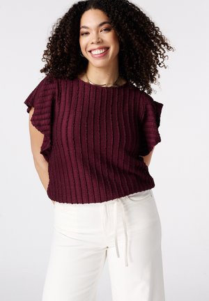 Lachende vrouw met krullend haar, gekleed in een gestructureerde donkerpaarse top met korte mouwen en witte broek met trekkoord tegen een witte achtergrond.