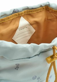 Svetlomodra vrečka z vezalkami z notranjostjo v bež barvi, z obeskom za ime. Zunanjost ima subtilne okrogle vzorce in trpežno teksturo blaga.