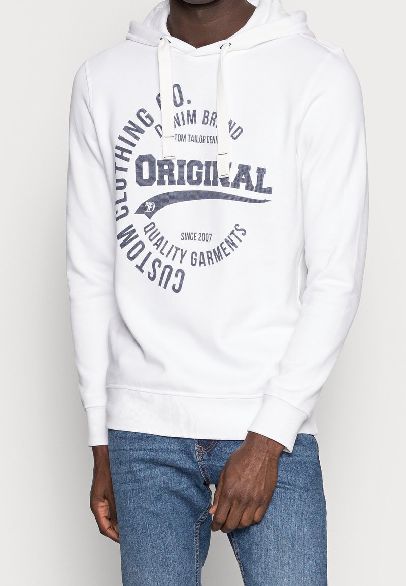 Homme portant un sweat à capuche blanc avec un texte bleu marine "Original Custom Clothing Co. Quality Garments" et un jean bleu sur un fond uni.