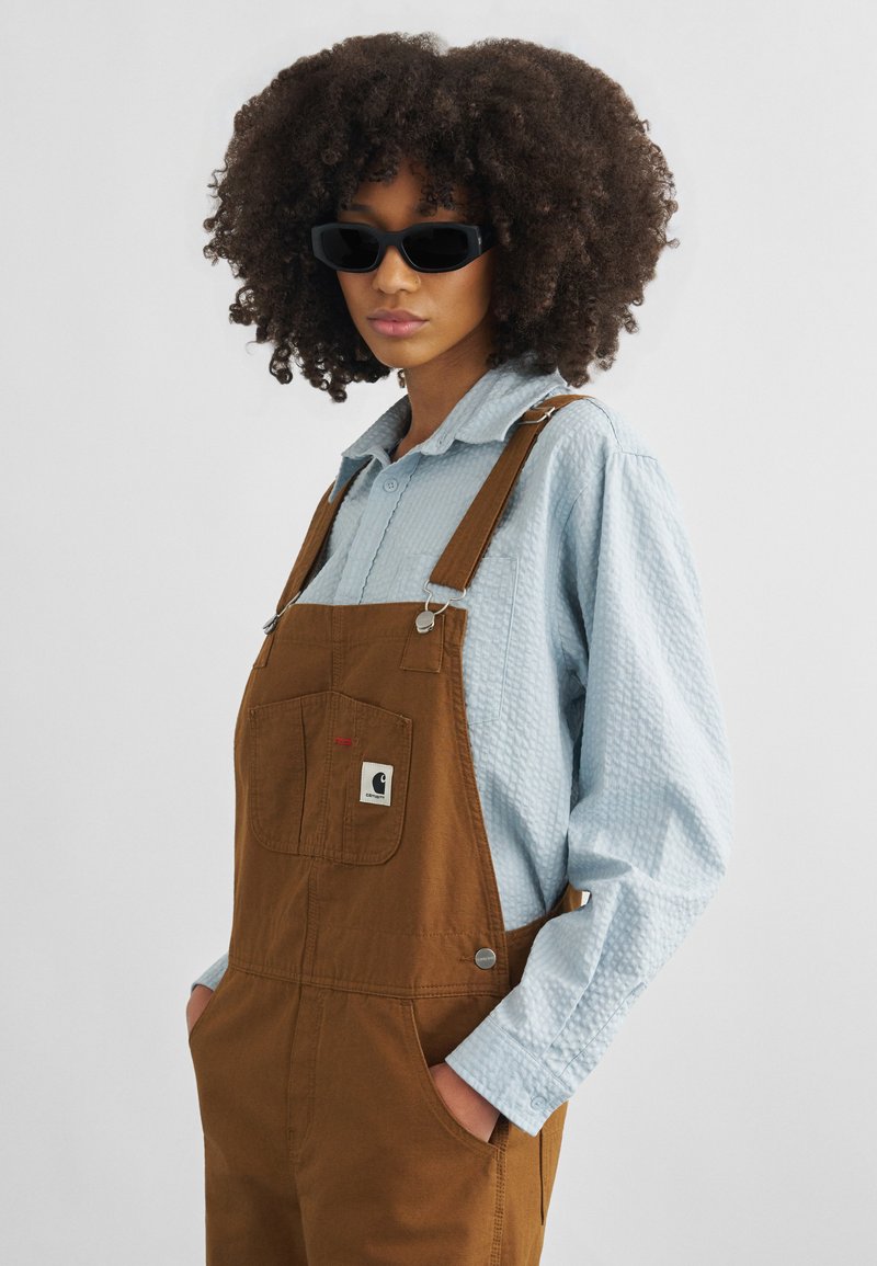 Young person with curly hair wearing black sunglasses, a light blue textured shirt, and brown overalls with hands in pockets.