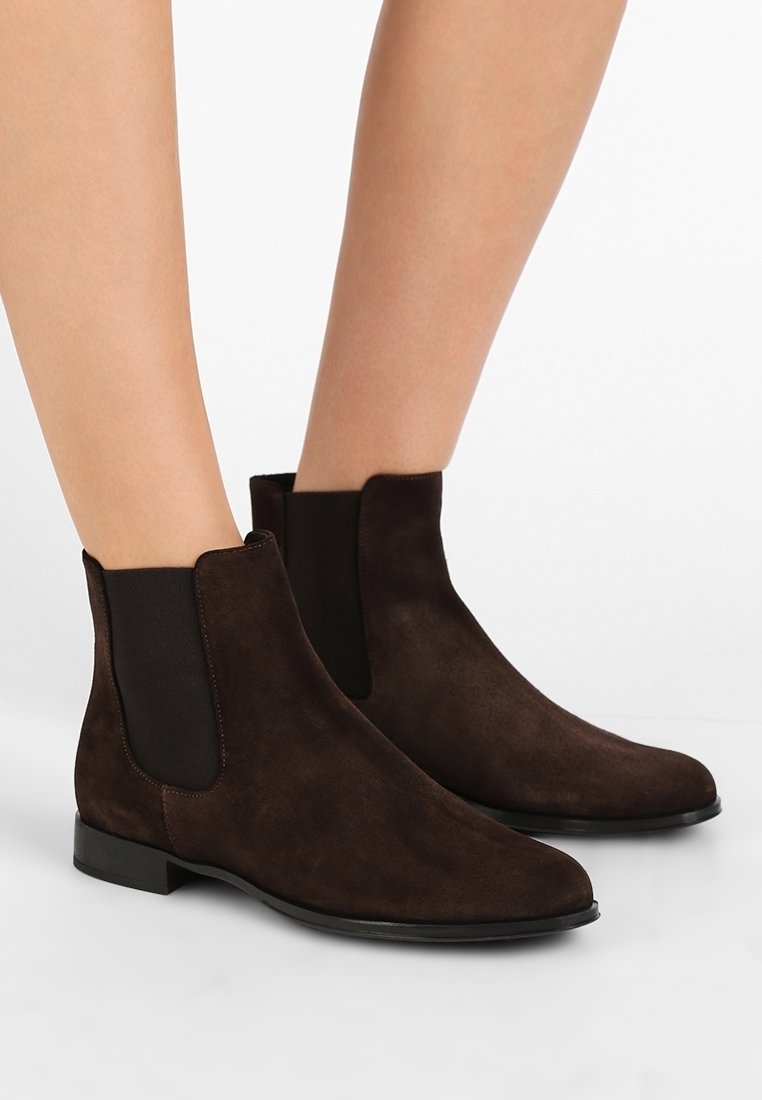 Bottines Chelsea en suède marron foncé montant à la cheville avec des panneaux latéraux élastiques, portées sur jambes nues sur un fond uni.