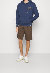 Uomo in felpa con cappuccio blu navy con scritta frontale, pantaloni corti marroni e sneakers bianche, in piedi su uno sfondo chiaro e semplice.
