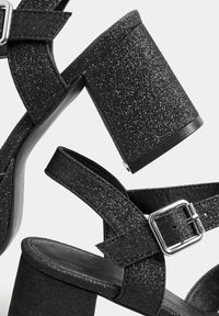 Sandalias de tacón alto con glitter negro y un tacón grueso, correa texturizada y detalle de hebilla ajustable en plata. Plantilla suave y materiales sintéticos.