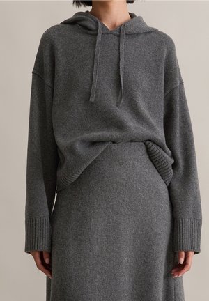 Personne portant un sweat à capuche en maille gris foncé avec des cordons, assorti à une jupe en maille taille haute, sur un fond neutre.