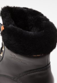 Stivale in pelle nera con un morbido risvolto in pelliccia nera, dotato di occhielli dorati e dettagli di lacci marroni a contrasto vicino alla caviglia.