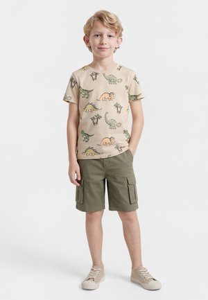Blonder Junge steht mit den Händen in den Taschen, trägt ein beiges T-Shirt mit Dinosaurierdruck, olivfarbene Cargo-Shorts und beigen Sneakers vor weißem Hintergrund.