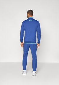 Blauer Trainingsanzug aus leichtem Material, mit einem Kragen mit kontrastierendem Besatz und einem Logo auf dem oberen Rücken. Die Hose hat schmal zulaufende Beine.