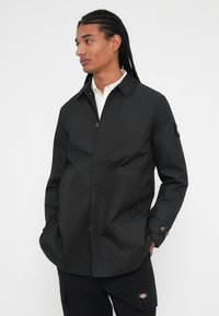 Giacca leggera nera con colletto, bottoni frontali e tasche laterali. Tessuto liscio, con un logo discreto sulla manica.