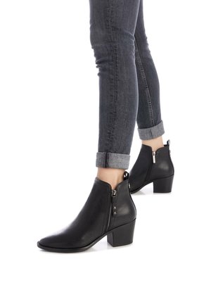 Botas de cuero negras hasta el tobillo con punta afilada, cierre de cremallera y tacón apilado. Llevadas con jeans oscuros remangados. Textura suave.