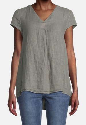 Vrouw draagt een loszittende, korte mouw, grijze linnen top met V-hals, gecombineerd met een blauwe spijkerbroek, staand tegen een effen achtergrond.