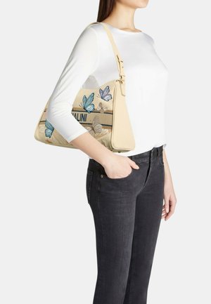 Donna con top bianco e jeans neri tiene una borsa a tracolla beige con disegni di farfalle blu e marroni.