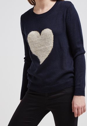 Marineblauwe trui met een groot, pluizig beige hart aan de voorkant, gedragen met zwarte broek.