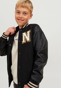 Svart varsityjacka med lädersleeves, med en krämfärgad och gyllene bokstav "N"-patch. Ribbad mudd och midja med krämfärgade ränder.