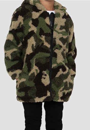 Enfant portant une veste polaire camouflage verte, marron et beige, avec une fermeture éclair à l'avant et les mains dans les poches.