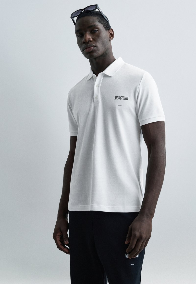 Νέος άνδρας που φοράει λευκό πόλο Moschino και μαύρο παντελόνι Moschino, με γυαλιά ηλίου τοποθετημένα στο κεφάλι, στέκεται μπροστά σε απλό φόντο.