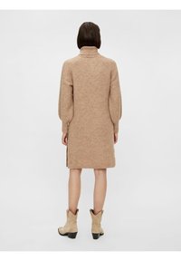 Pletené béžové šaty ke kolenům s vysokým límcem, žebrovanou texturou, dlouhými rukávy a postranními rozparky. V kombinaci se světle béžovými kotníkovými botami.