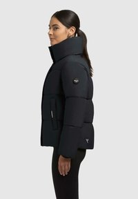 Donna con capelli scuri raccolti in una coda di cavallo, che indossa un giubbotto imbottito nero con colletto alto, in piedi di profilo su uno sfondo chiaro.